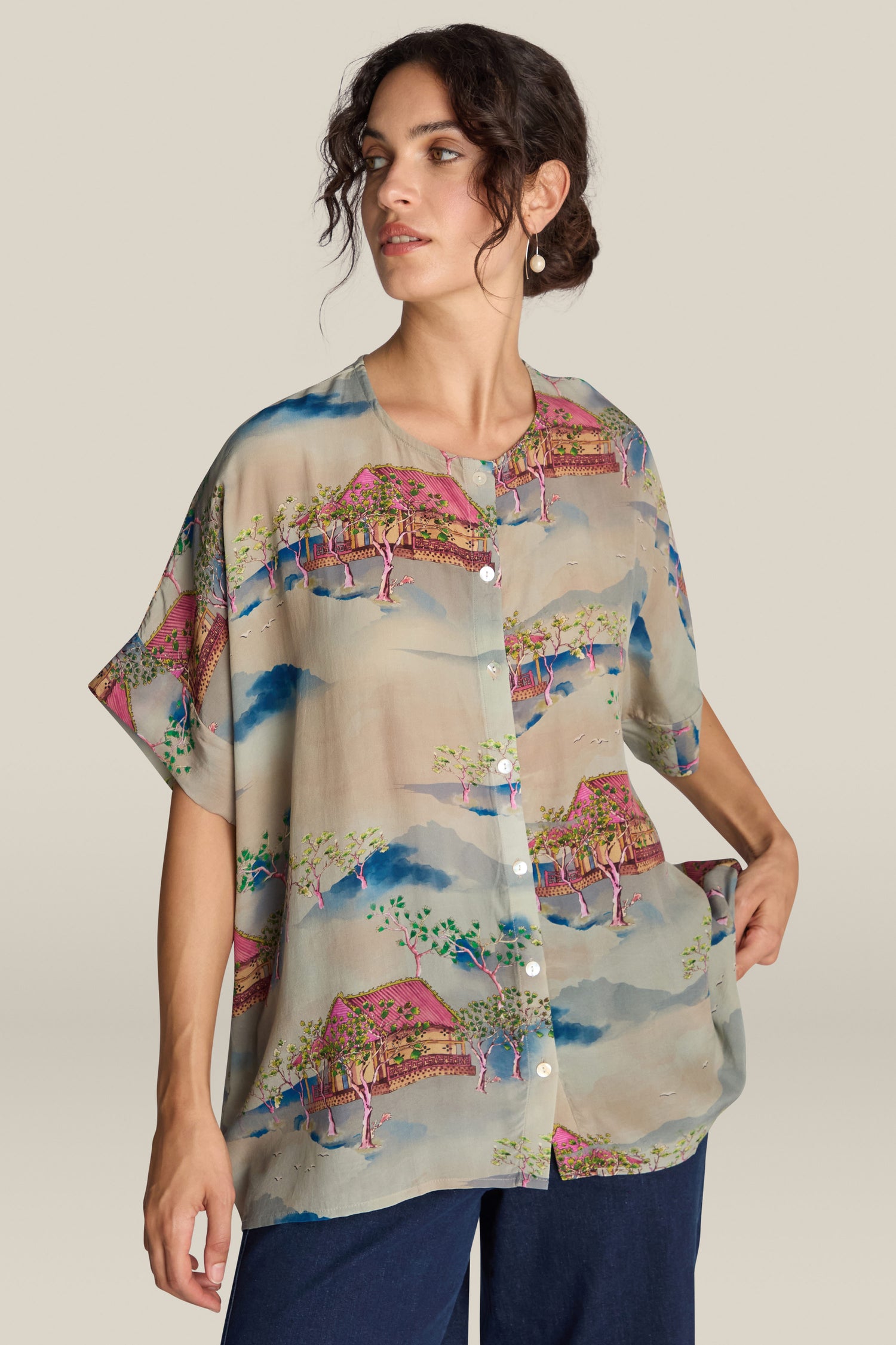 A person is wearing the Landscape Blouse, a loose, short-sleeved, button-up shirt adorned with a vibrant landscape pattern reminiscent of Japanese watercolour paintings featuring trees and huts. They have one hand in their pocket and are looking to the side.