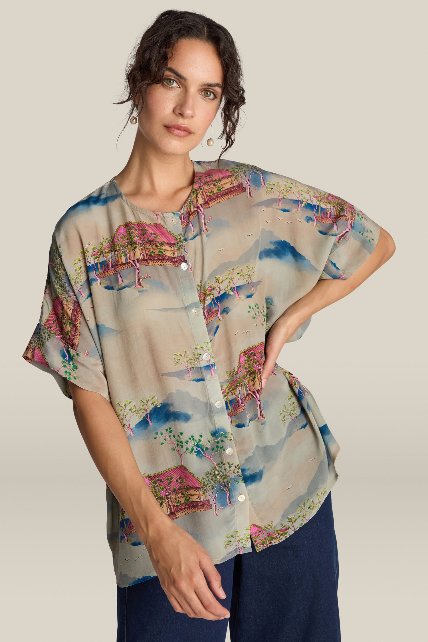 A woman with dark hair is wearing a Landscape Blouse featuring a beautiful pattern, paired with blue pants. Crafted from a soft Viscose blend, the blouse feels like wearable art. She poses with one hand on her hip against a plain background.