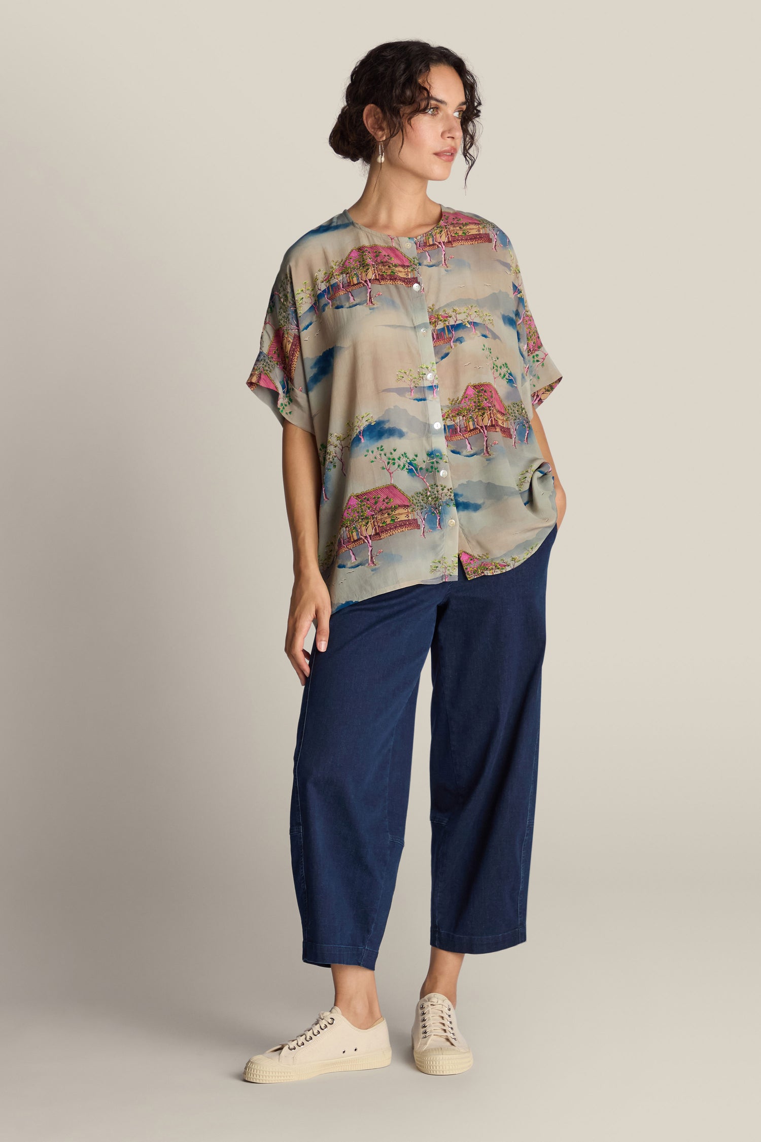 A person wearing the Landscape Blouse, a colorful, patterned button-up shirt resembling wearable works of art, along with blue wide-leg pants and white sneakers, stands against a plain background with hands in pockets.