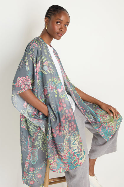 A woman sits on a stool wearing a white top, light gray pants, and the Boho Garden Print Throwover in shades of blue, pink, and green.