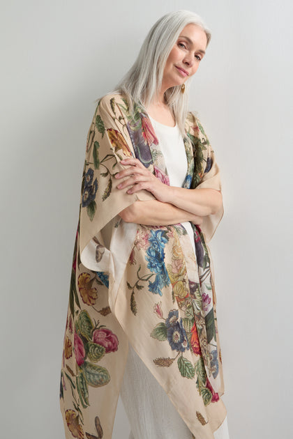 Woman with long gray hair in a cream outfit and the Primula Throwover, a bold floral kimono, stands with arms crossed against a plain light background.