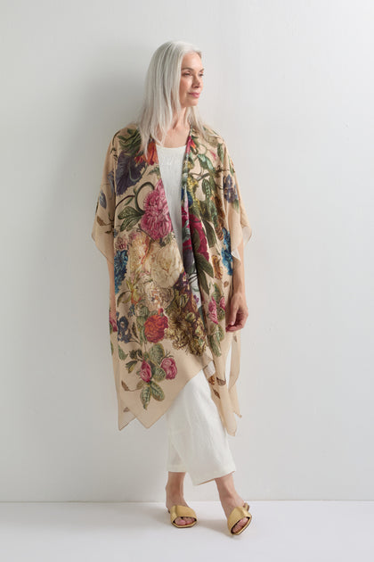 A woman with long gray hair wears the Primula Throwover, a beige floral kimono, over a white outfit and yellow sandals, standing against a plain white wall.
