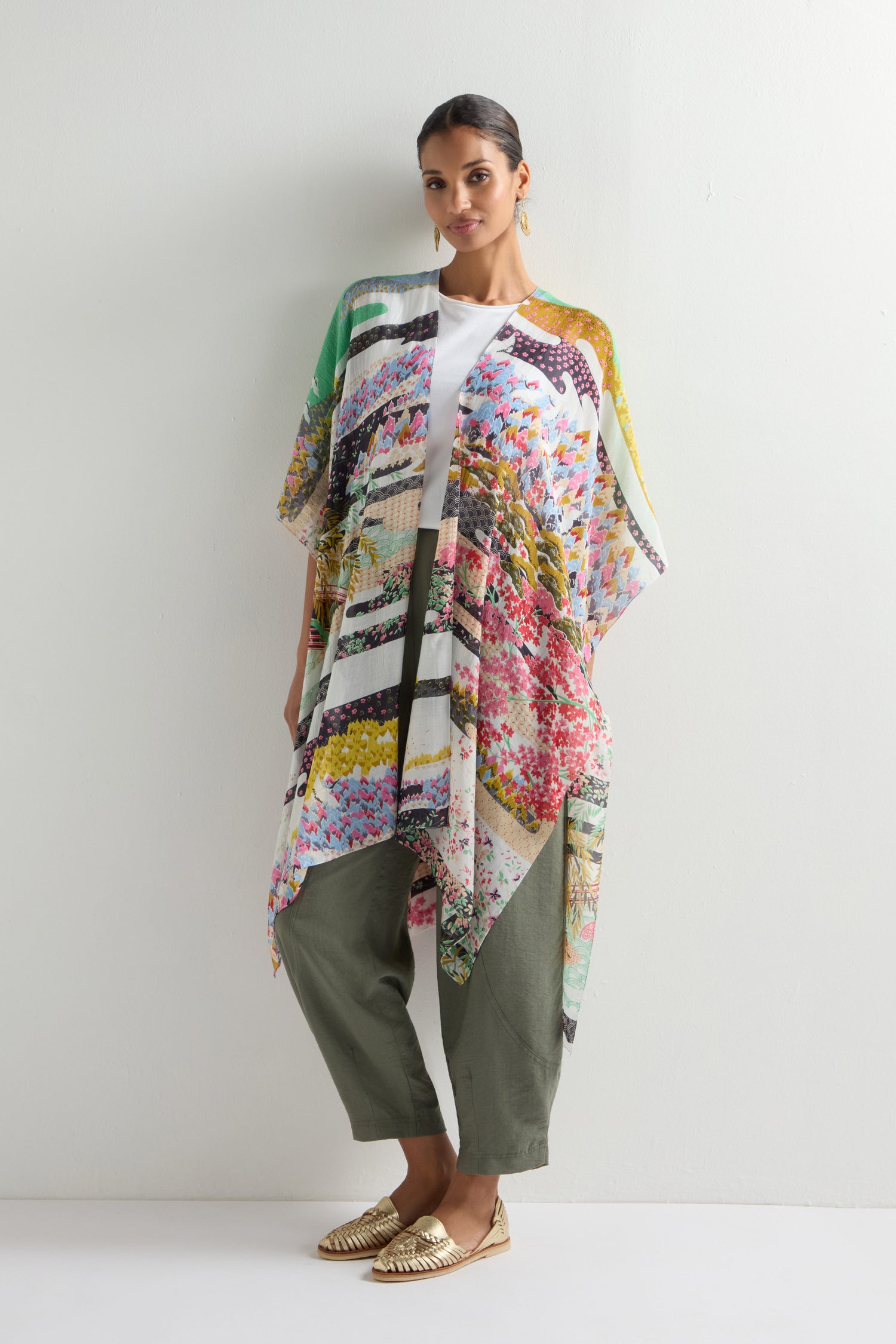 A person wearing a white shirt and olive pants dons the Spring Print Throwover, a colorful, patterned cover-up inspired by Japanese landscapes, paired with gold shoes against a plain light wall.