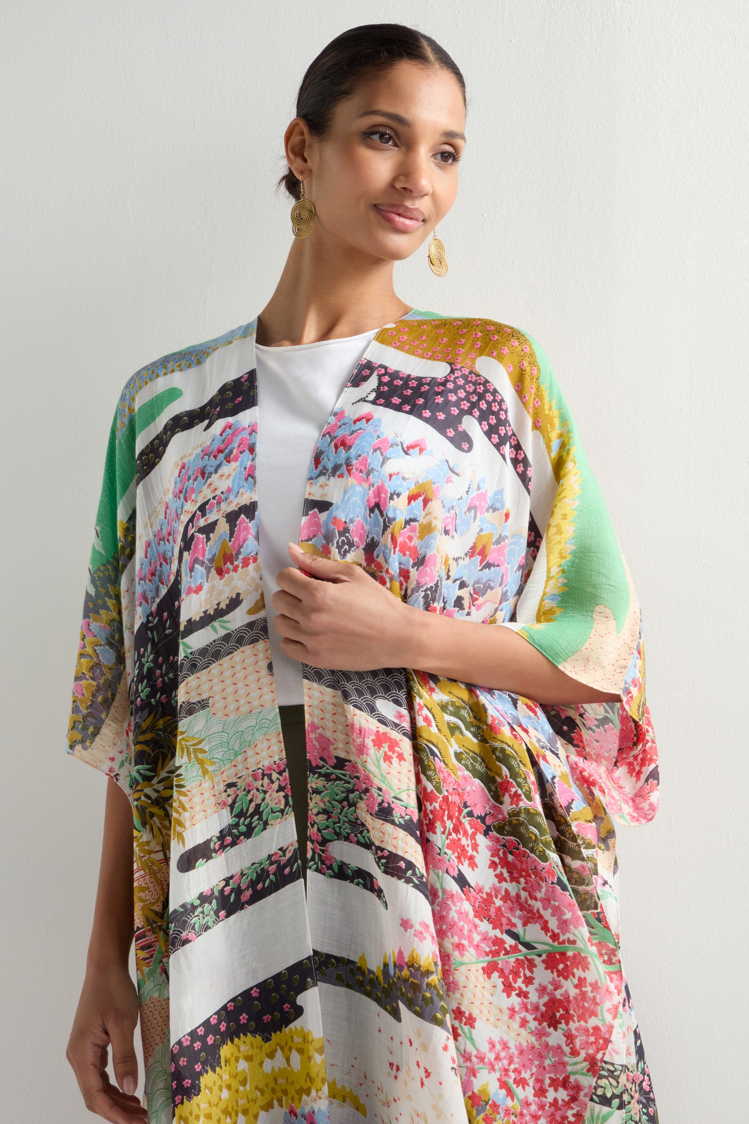 Wearing a colorful Spring Print Throwover, inspired by Japanese landscapes, a person stands against a plain background. This kimono-style jacket adds elegance to the white top.