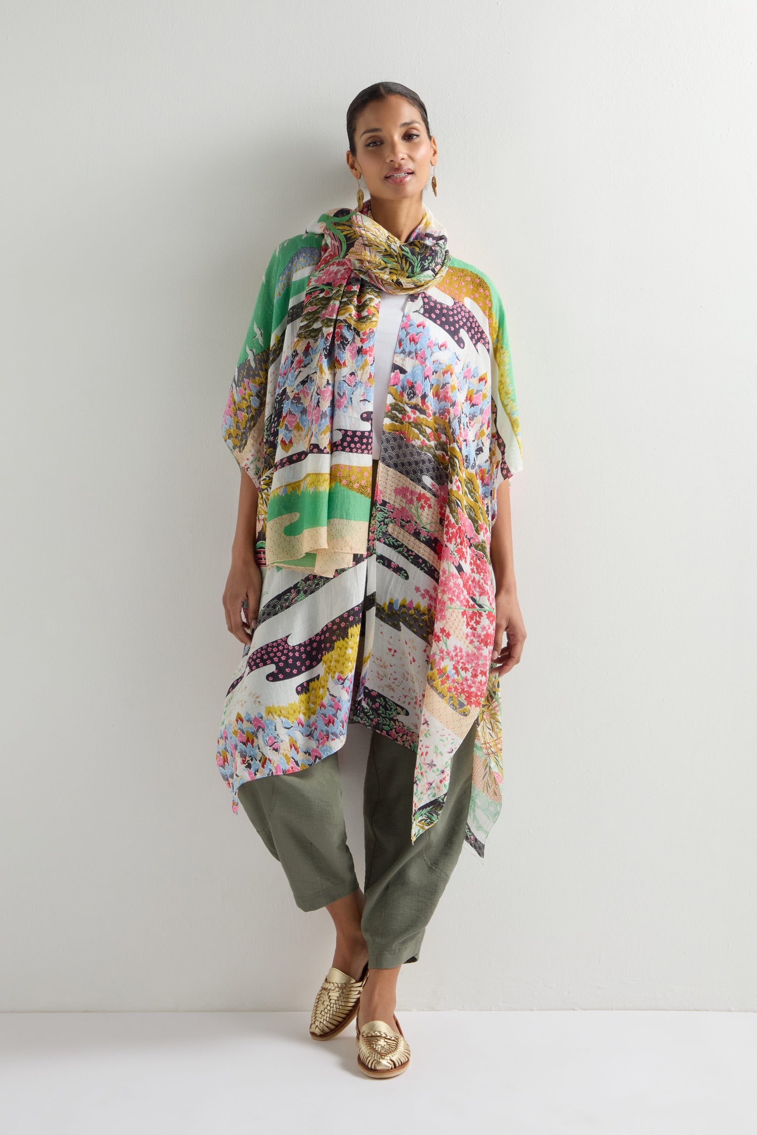 A person stands against a white wall, draped in the Spring Print Throwover, featuring colorful patchwork reminiscent of oriental garden designs, enhancing their white top and green pants.