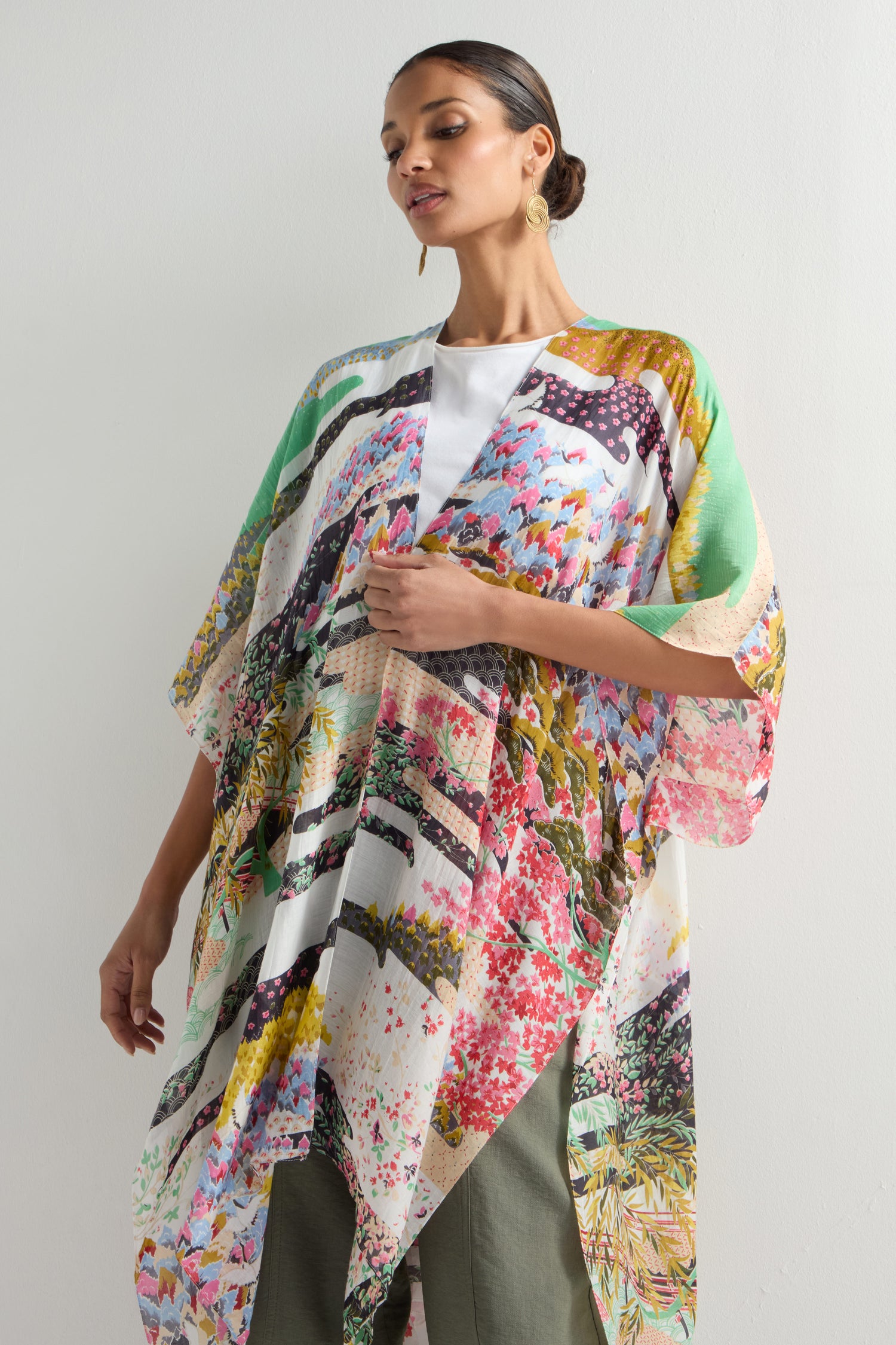 A woman elegantly wears the Spring Print Throwover, a colorful kimono-style robe inspired by Japanese landscapes, over a white top and green pants against a plain white backdrop.