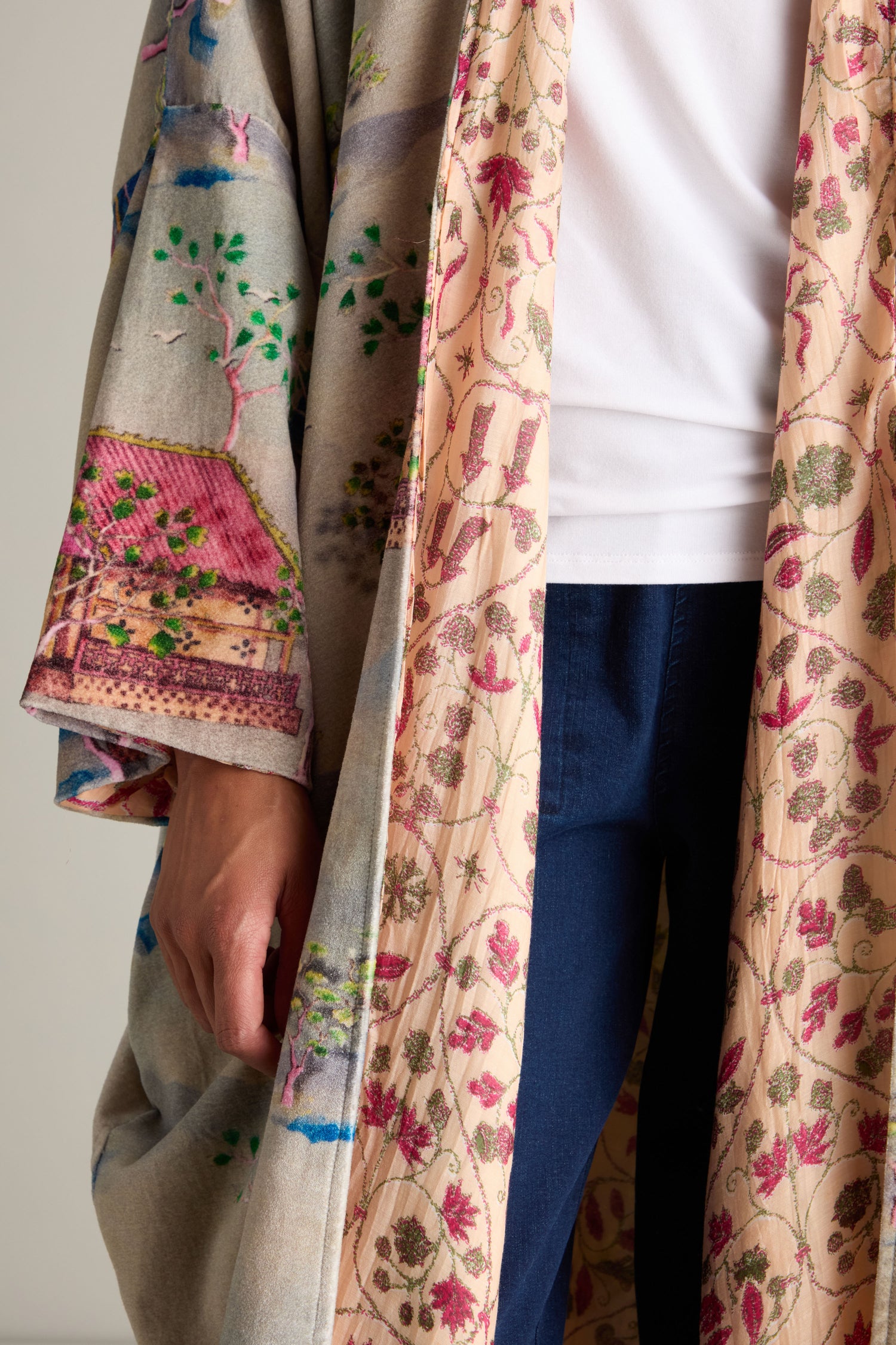 A person is wearing a white shirt and dark blue pants, topped with the luxurious Landscape Velvet Coat. This multicolored, patterned robe features intricate designs and scenes reminiscent of a Japanese landscape print.