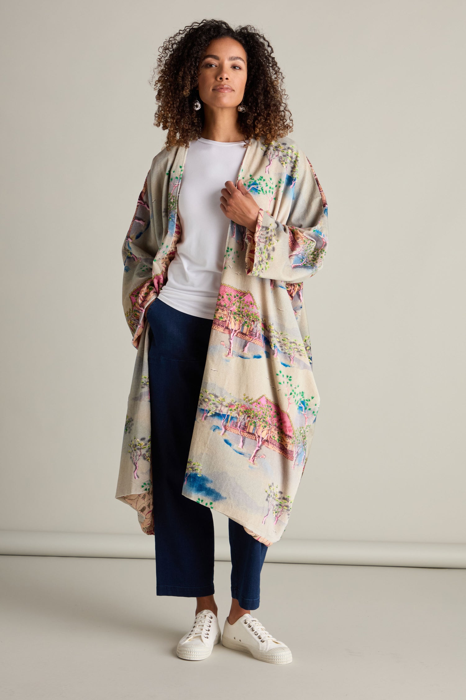 A woman with curly hair stands against a neutral backdrop, wearing the luxurious Landscape Velvet Coat featuring a Japanese landscape print over a white top and dark pants, paired with white sneakers.