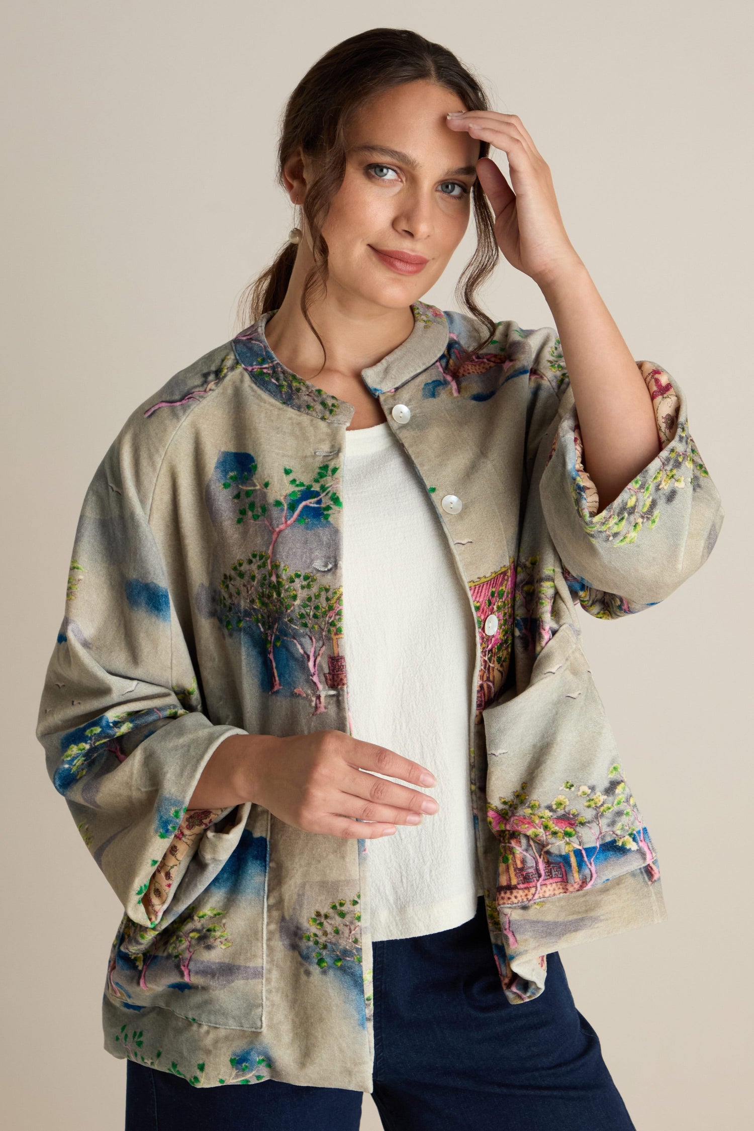 A person wearing the Landscape Velvet Jacket, featuring wide raglan sleeves and a colorful patterned design, over a white shirt poses against a plain background. They have one hand on the jacket and the other near their head.