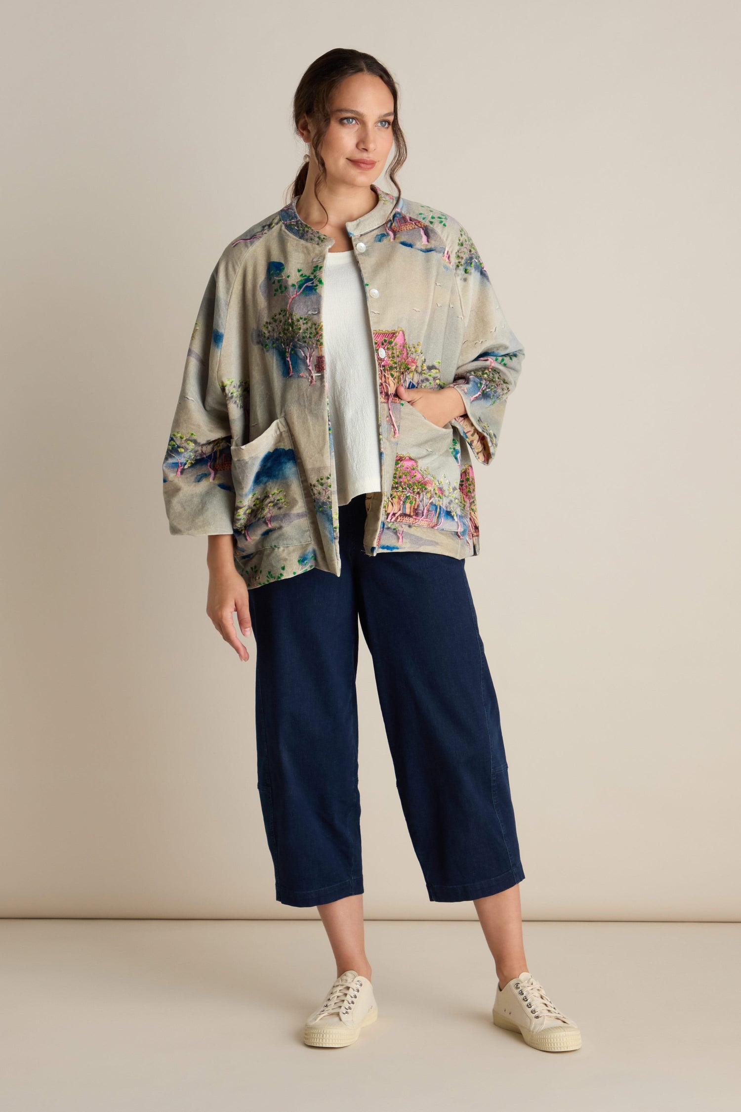 A woman stands against a plain background wearing the Landscape Velvet Jacket, which features a light-colored patterned design with wide raglan sleeves. She pairs it with a white shirt, dark blue cropped pants, and white sneakers while looking slightly to the side.