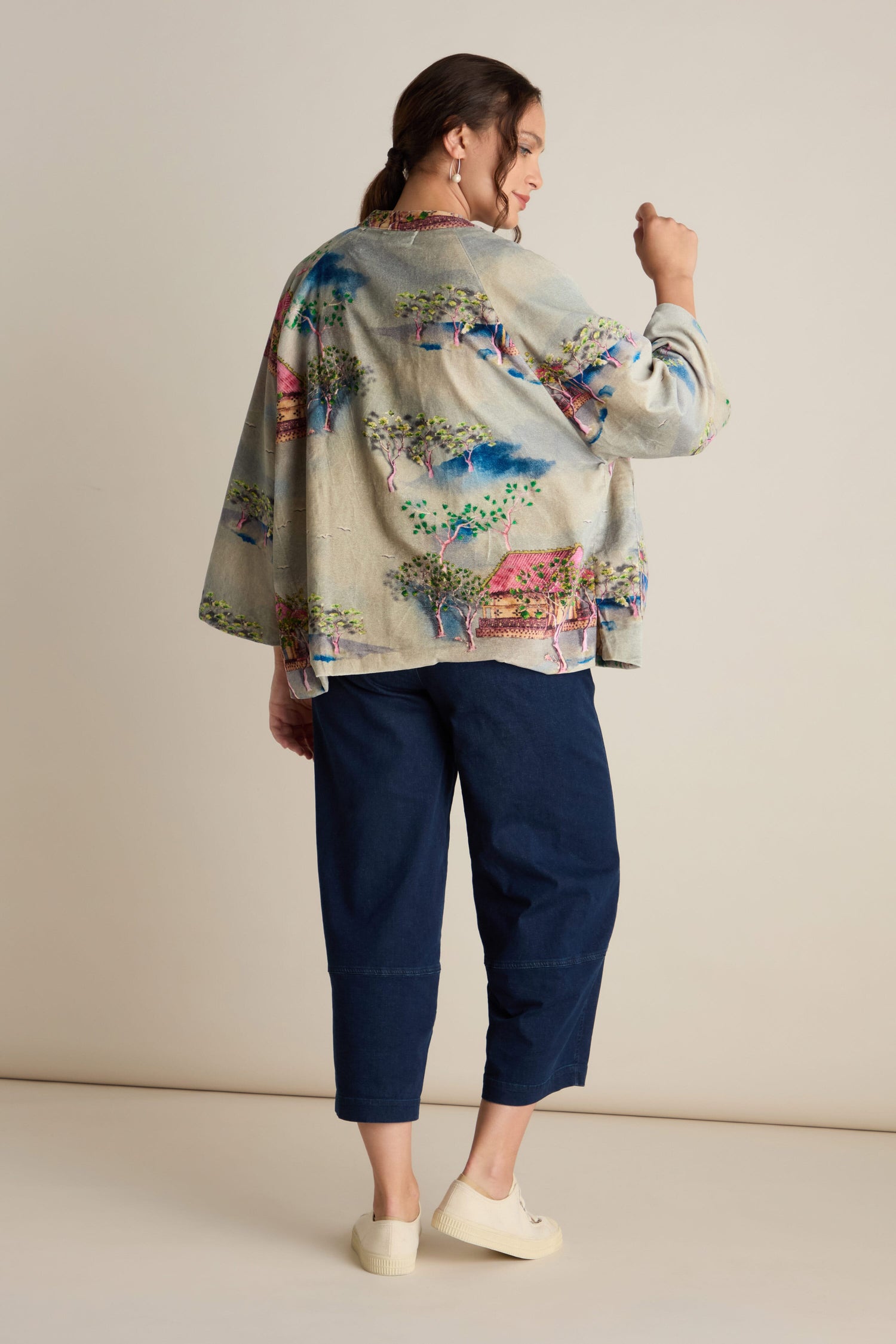 Person wearing a Landscape Velvet Jacket featuring intricate Japanese landscape watercolors, accompanied by dark blue pants and white shoes, standing with their back to the camera. The jacket's colorful print and wide raglan sleeves add a touch of elegance to the ensemble.