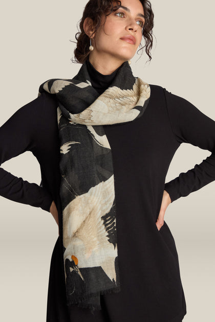 A person wearing a black long-sleeve top and a vintage-inspired Stork Print Woollen Scarf, featuring a luxury black and white bird design that blends timeless elegance with contemporary flair.