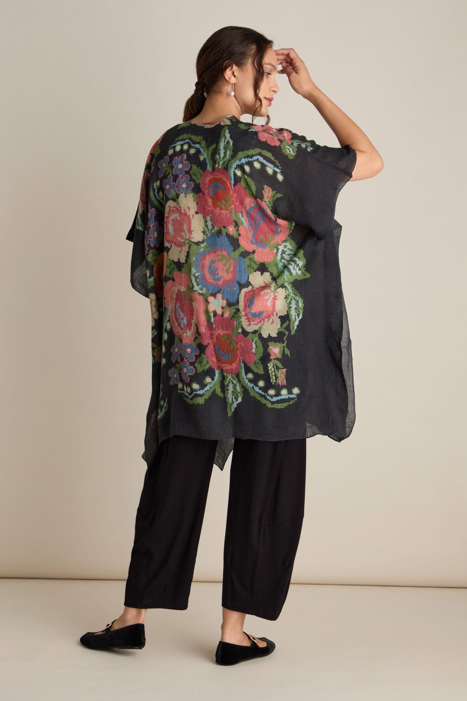 A person stands sideways, wearing black pants and a black top featuring the large colorful digital print floral pattern of the Woven Flower Wool Throwover on the back. They touch their head with one hand, showcasing this ideal layering piece for any season.