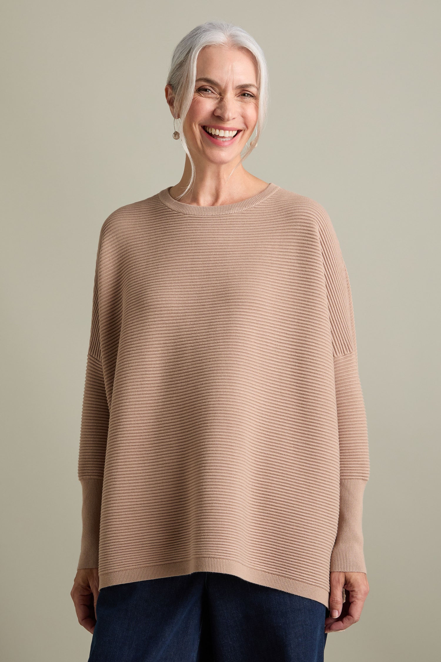 An older woman with long gray hair smiles while wearing a loose-fitting Ribbed Jumper in new-season colors. She stands against a plain background, exuding a luxurious feel.