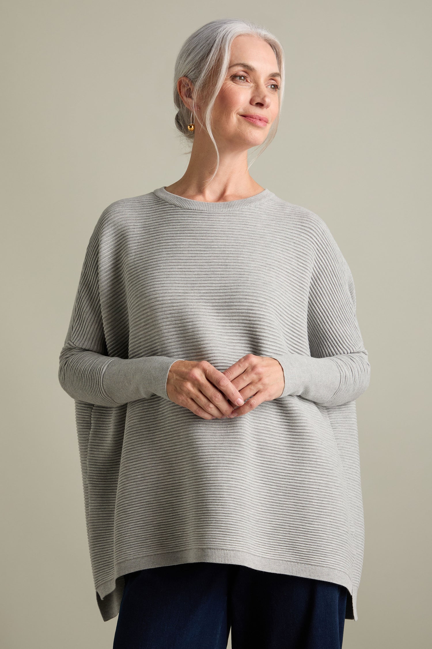 A woman with gray hair smiles while wearing a light gray Ribbed Jumper paired with dark pants, standing against a plain background. The luxurious feel of the jumper complements the new-season colors perfectly.