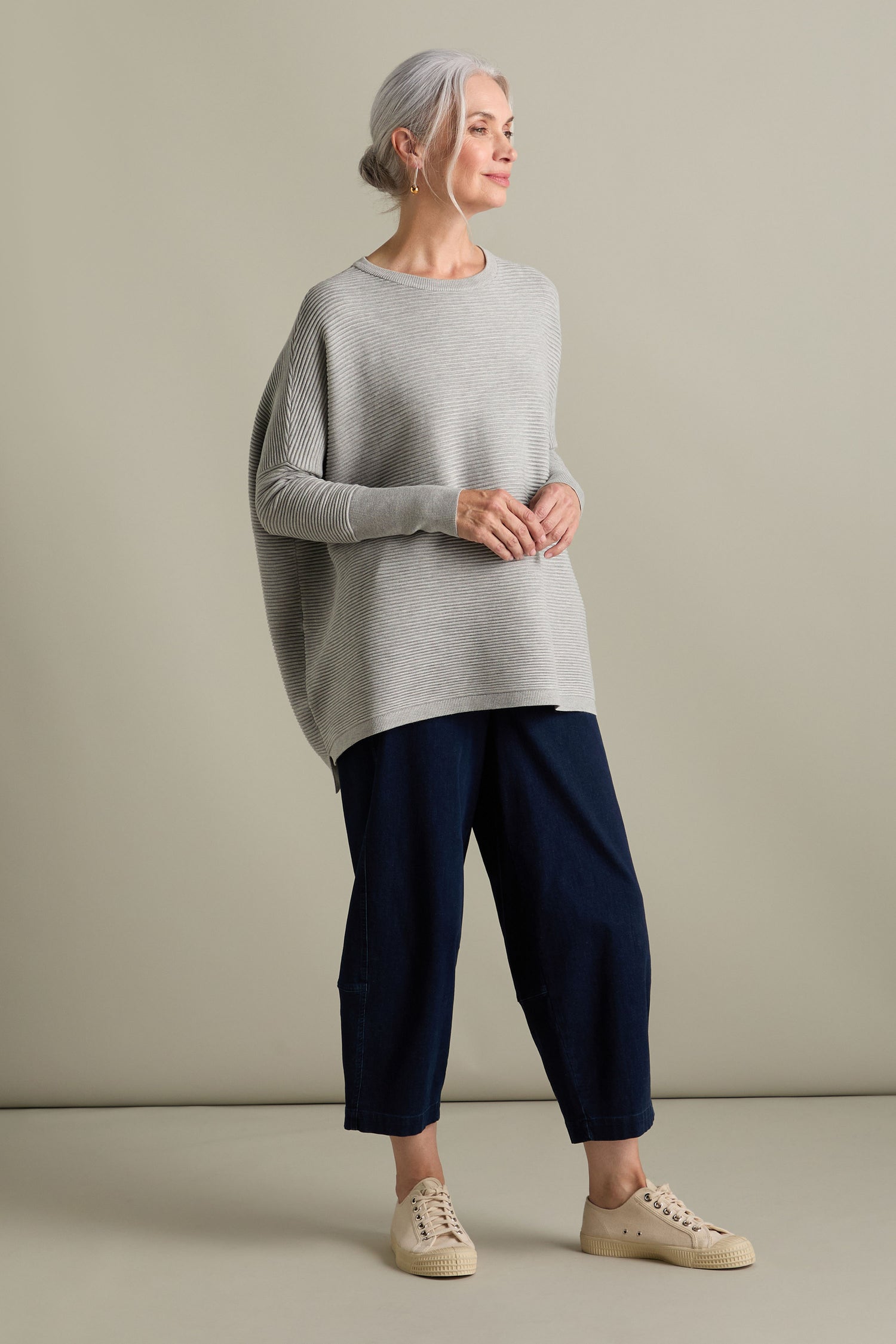 A woman with gray hair stands wearing a loose Ribbed Jumper from Paisie in new-season colors, along with dark blue pants and beige sneakers, against a plain background.