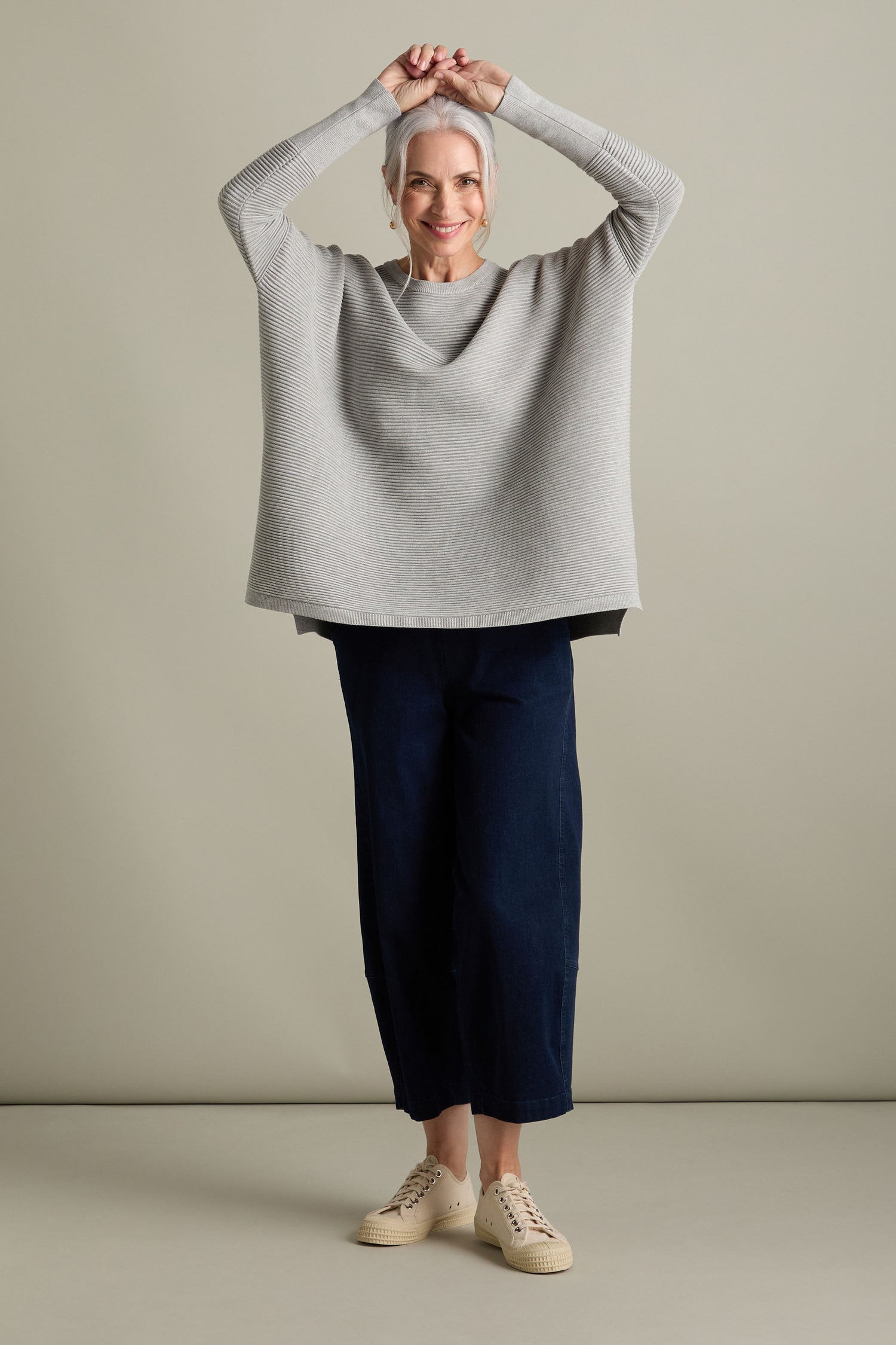 A person with gray hair is standing with arms raised, wearing a Ribbed Jumper in new-season colors, dark pants, and beige shoes against a neutral background.