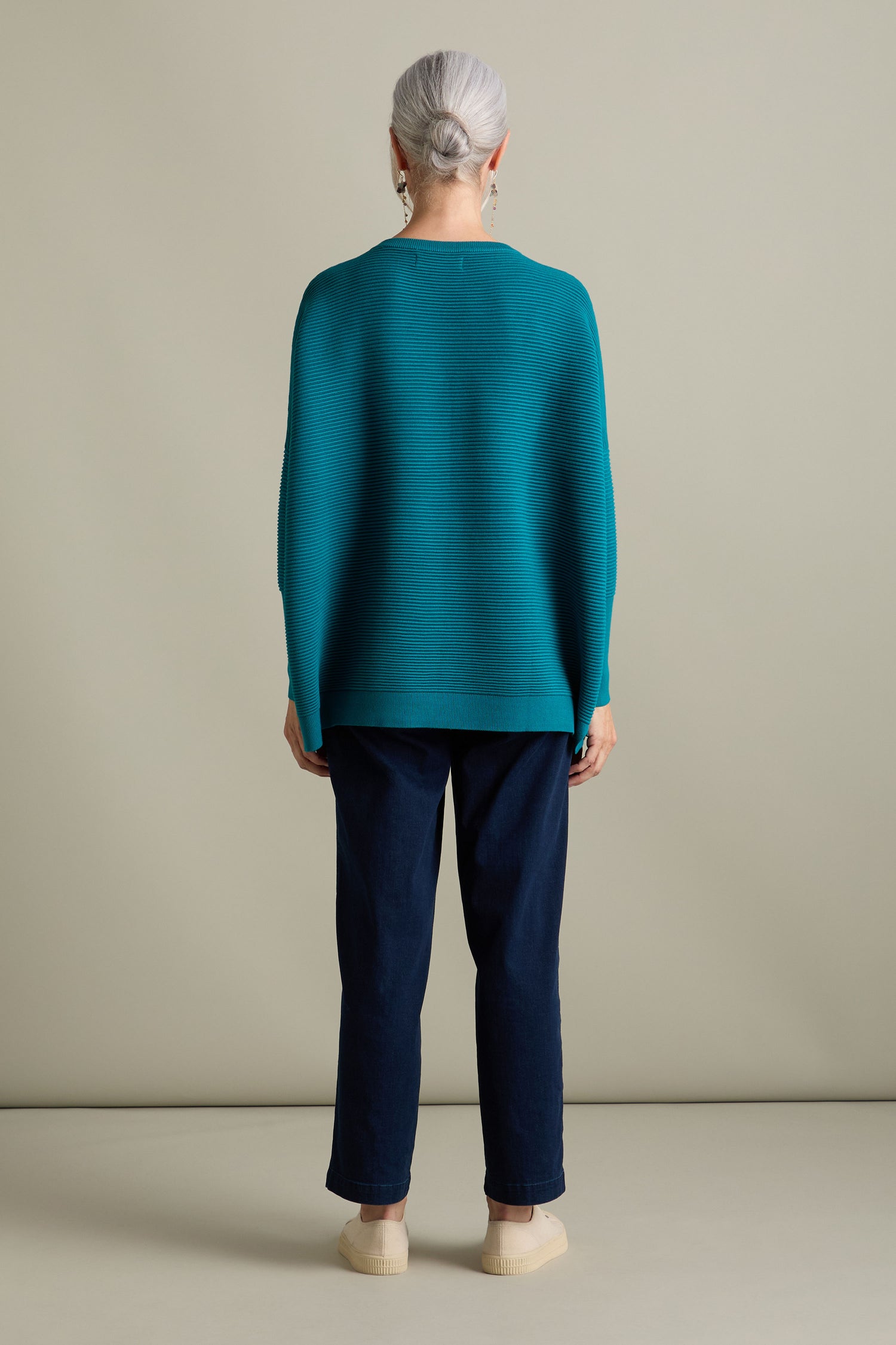 A person with gray hair, seen from behind, stands against a beige background wearing a teal Ribbed Jumper by Paisie, navy pants, beige shoes, and small earrings. This outfit embraces new-season colors while exuding a luxurious feel.