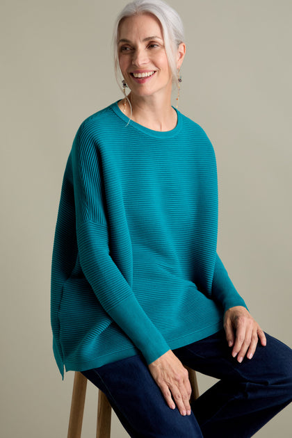 A woman with grey hair, wearing a Ribbed Jumper that exudes a luxurious feel in new-season colors, and dark jeans sits on a stool, smiling.