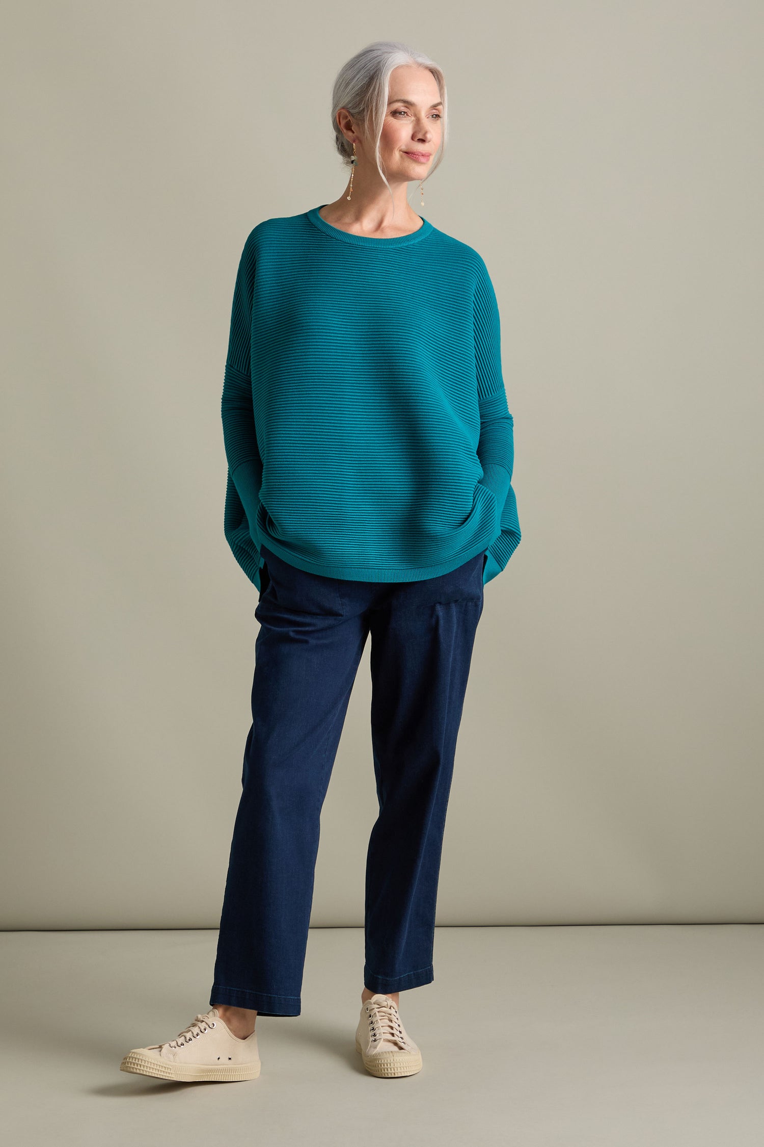 A person with grey hair, dressed in a Ribbed Jumper from Paisie's new-season collection, styled with dark blue pants and beige sneakers, stands with hands in pockets against a plain background, exuding a luxurious feel.