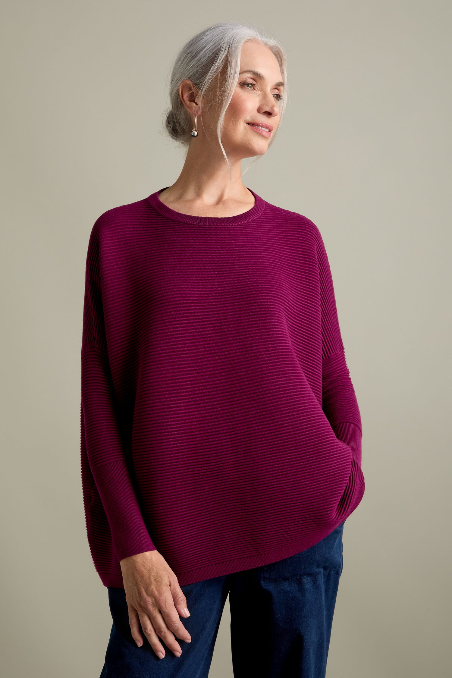 A woman with gray hair tied back is wearing a loose, new-season magenta Ribbed Jumper and dark pants, standing against a neutral background.