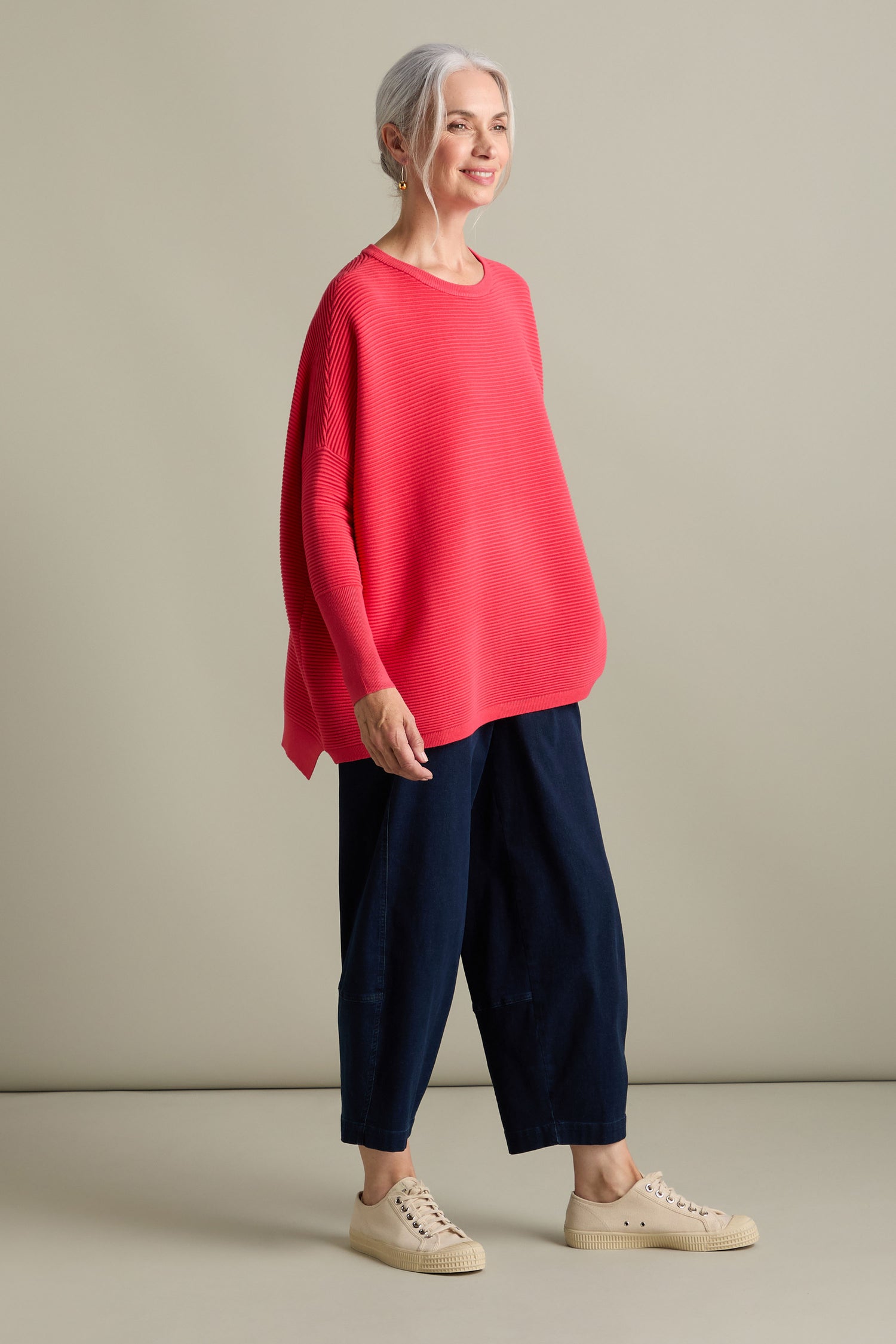 A woman with grey hair, wearing a Ribbed Jumper from Paisie's new-season collection and pairing it with navy blue pants and beige sneakers, stands and smiles against a plain background.