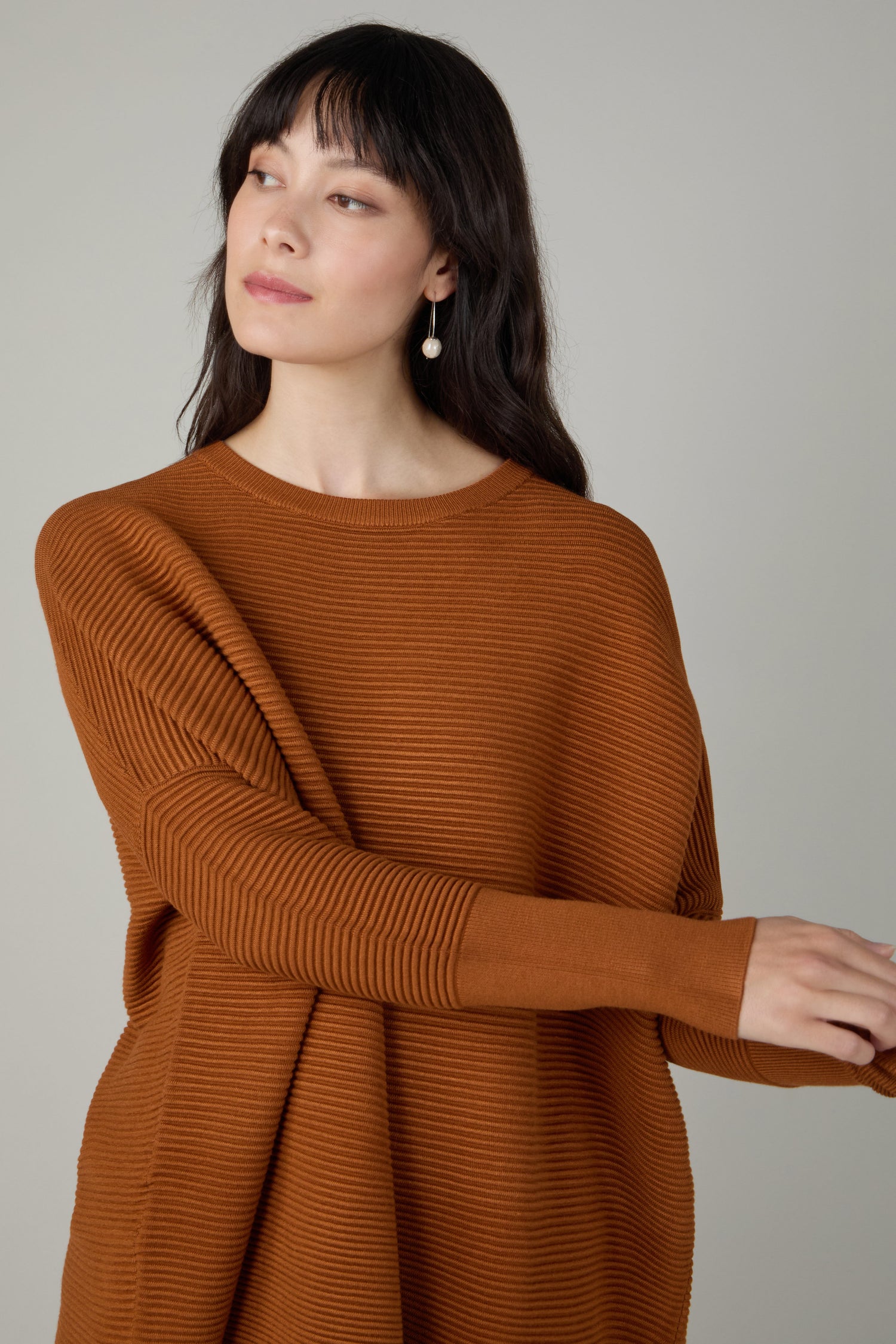Woman with long dark hair wears a Ribbed Jumper in rust, made from viscose-rich fabric, paired with pearl earrings. She looks to the side against a plain background.