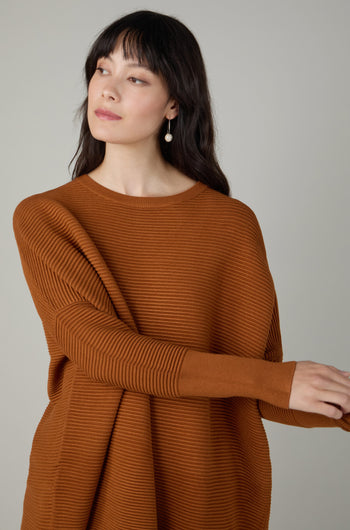 Woman with long dark hair wears a Ribbed Jumper in rust, made from viscose-rich fabric, paired with pearl earrings. She looks to the side against a plain background.