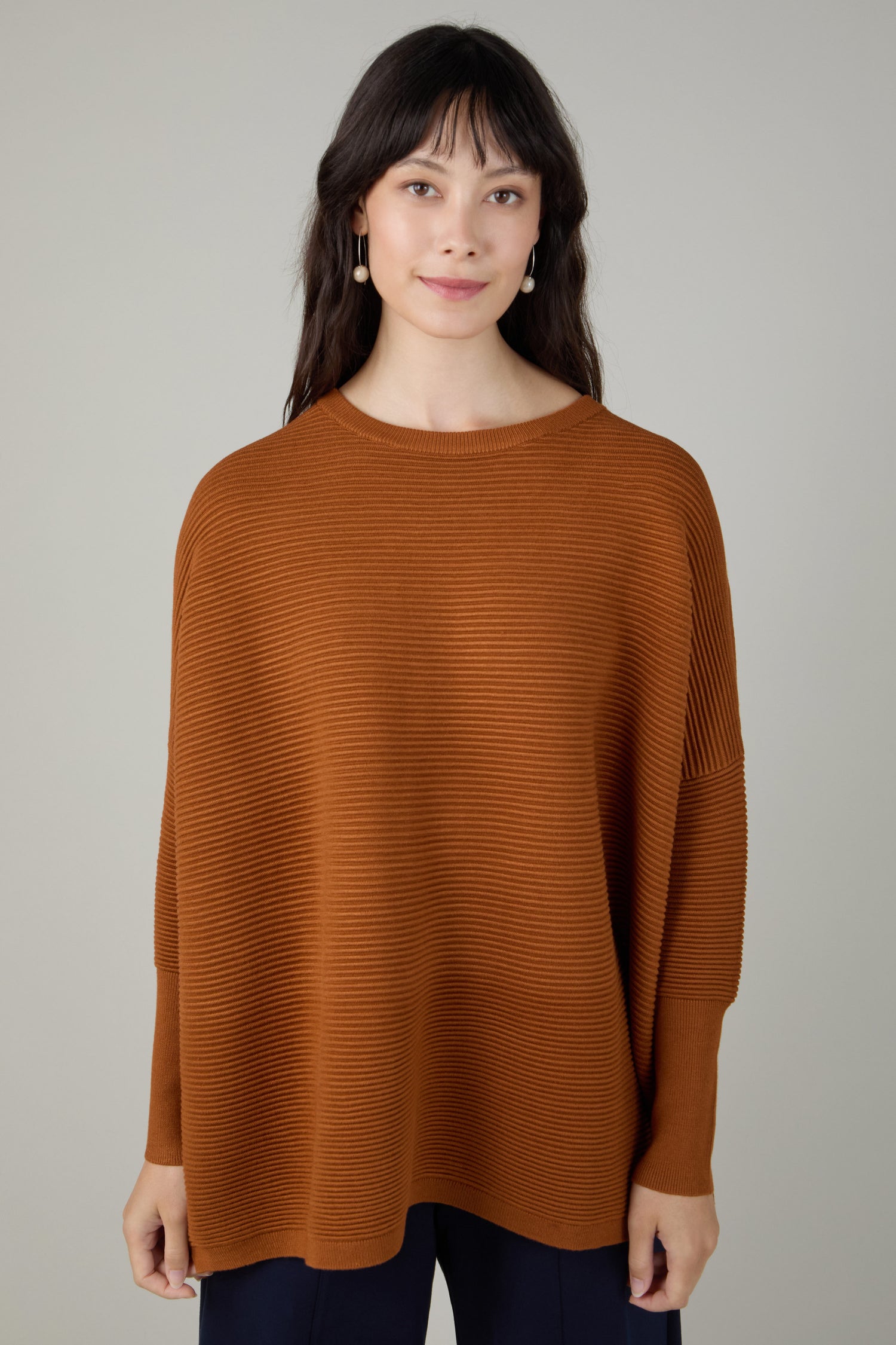 A woman with long dark hair wears a loose-fitting Ribbed Jumper by Paisie in rust, paired with navy pants, standing against a plain light gray background.