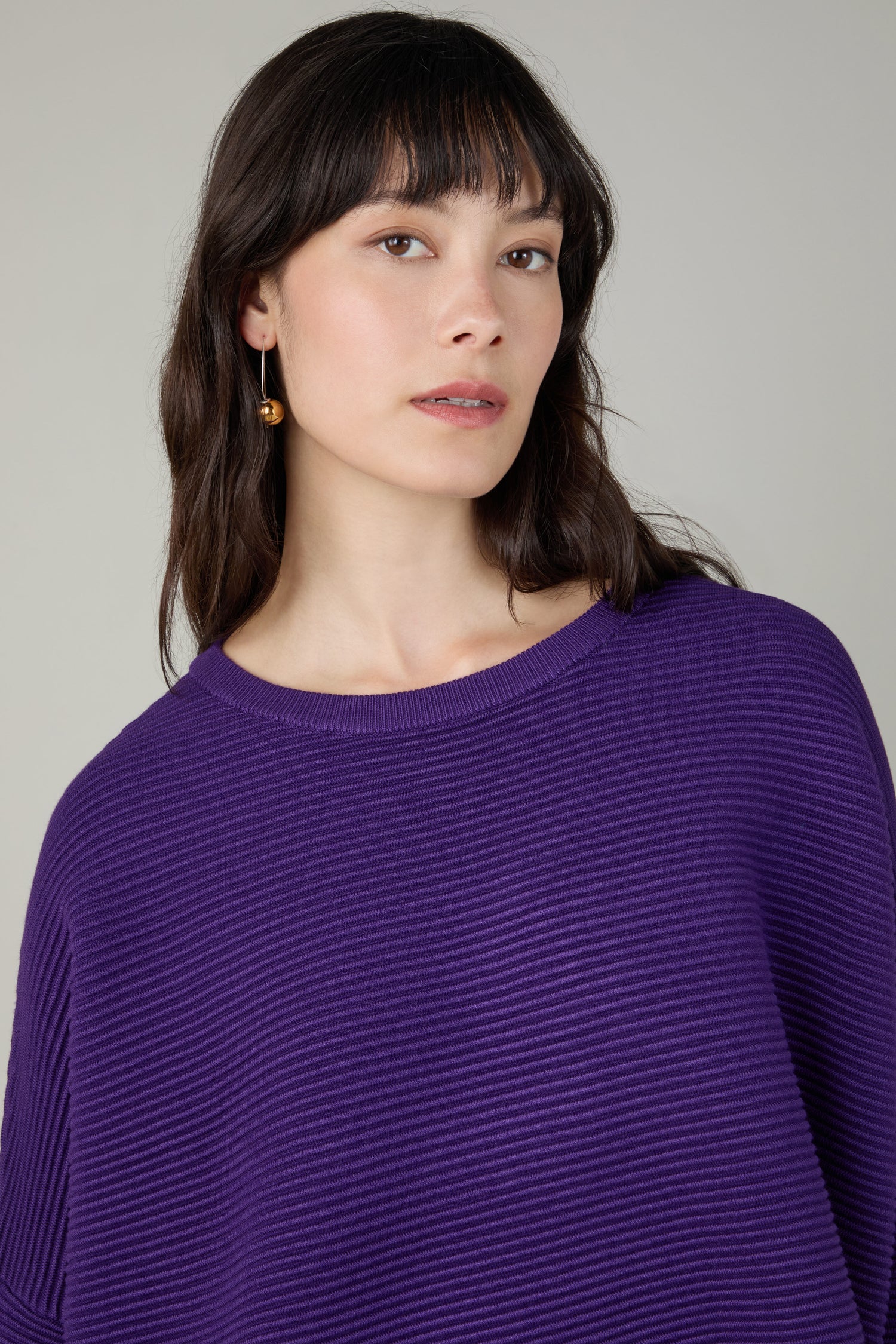 A woman with long dark hair and bangs wears a Ribbed Jumper in textured purple and a gold earring, looking slightly to the side against a neutral background.
