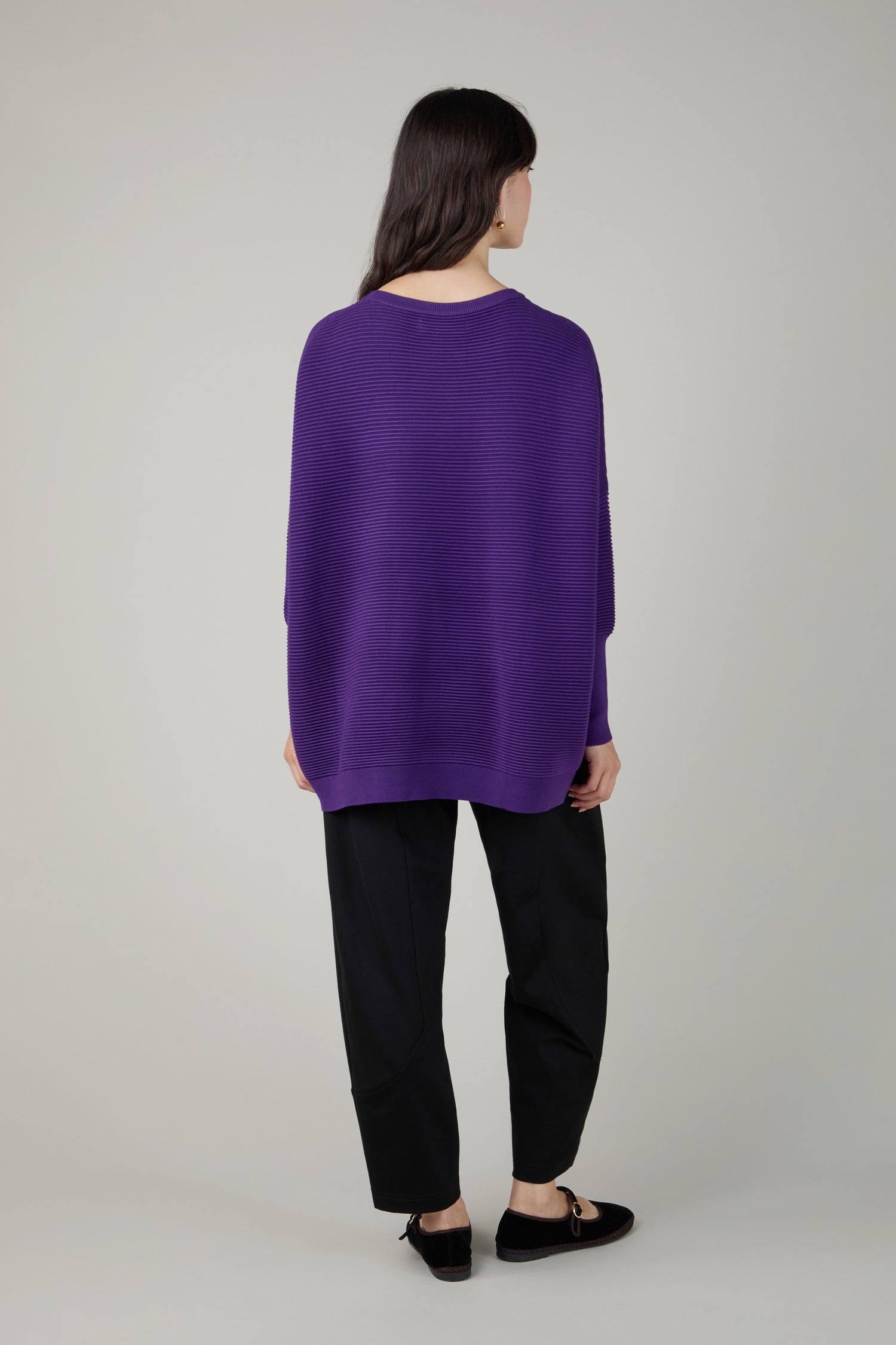 A person stands facing away, wearing a loose purple Ribbed Jumper with black pants and shoes against a plain light background.