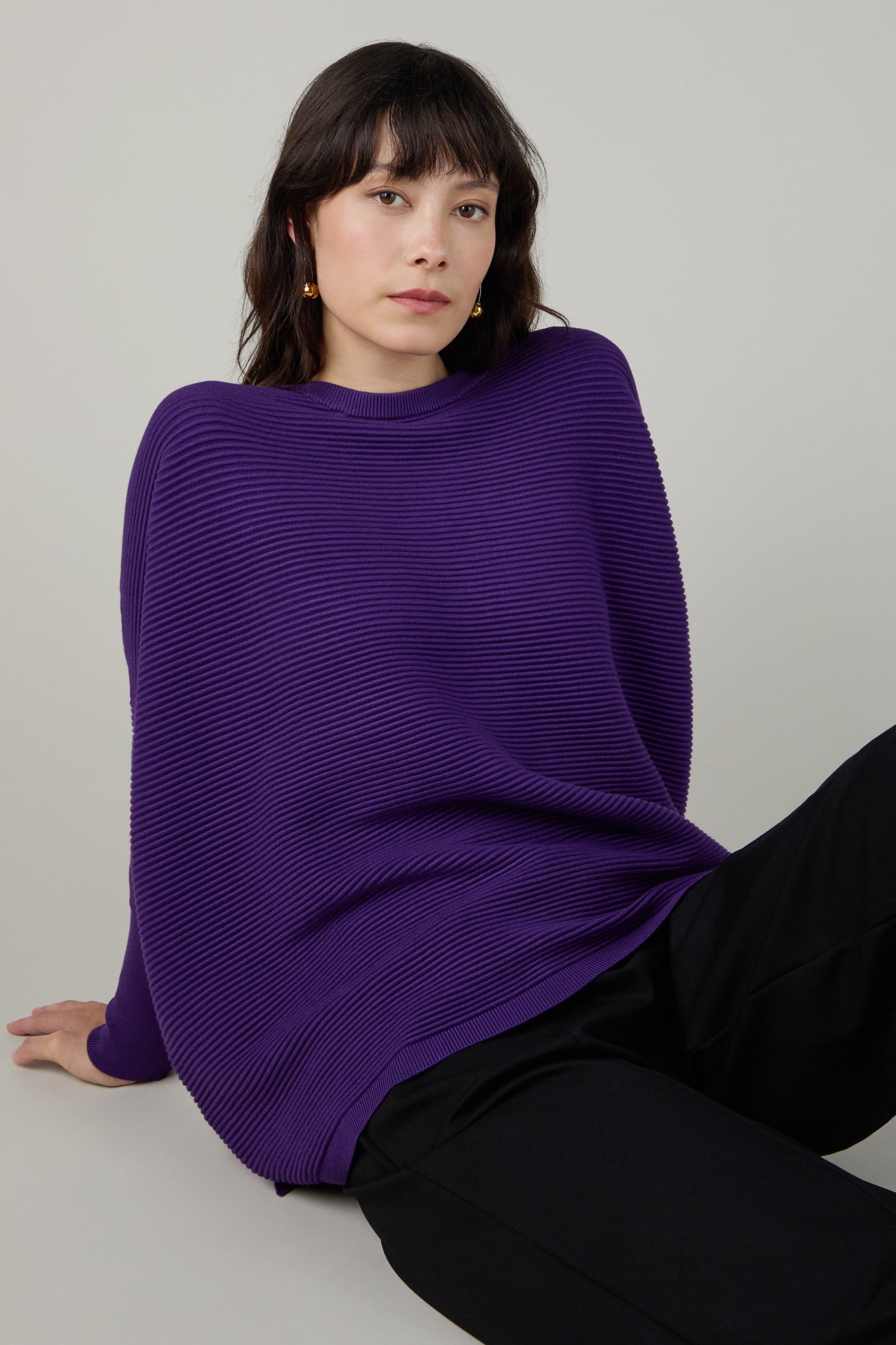 A person with dark hair sits on the floor wearing a purple Ribbed Jumper and black pants against a plain background.