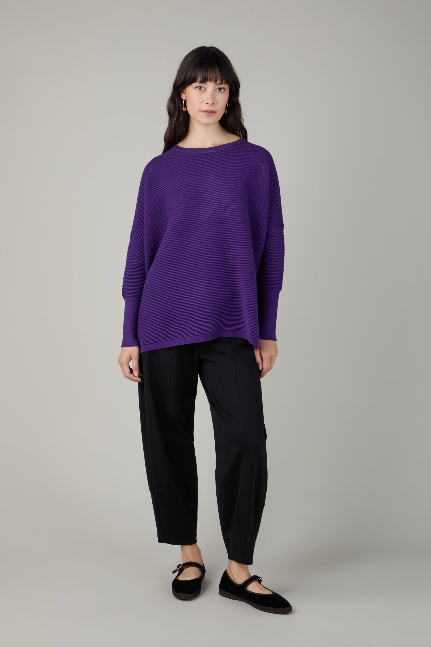 A woman stands facing forward wearing a Ribbed Jumper in purple, paired with black pants and black flat shoes, against a plain light gray background.