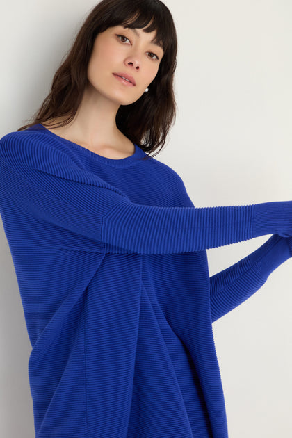 A woman with long dark hair wears the Round Neck Ribbed Top in bright blue, standing against a plain light background—a versatile piece for everyday layering.