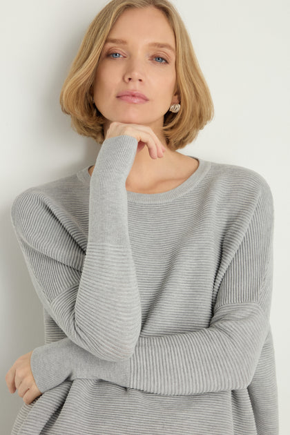 A woman with short blonde hair wears a light gray Round Neck Ribbed Top, standing against a plain background with one hand thoughtfully near her chin.