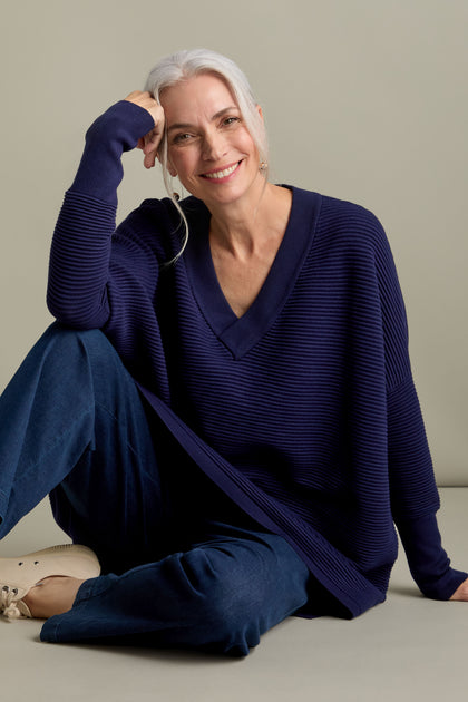 A woman with long gray hair smiles while wearing an oversized V Neck Ribbed Jumper and blue pants. Seated on the floor against a plain background, the luxurious ribbed knit design of her Paisie jumper adds a touch of elegance to her casual look.