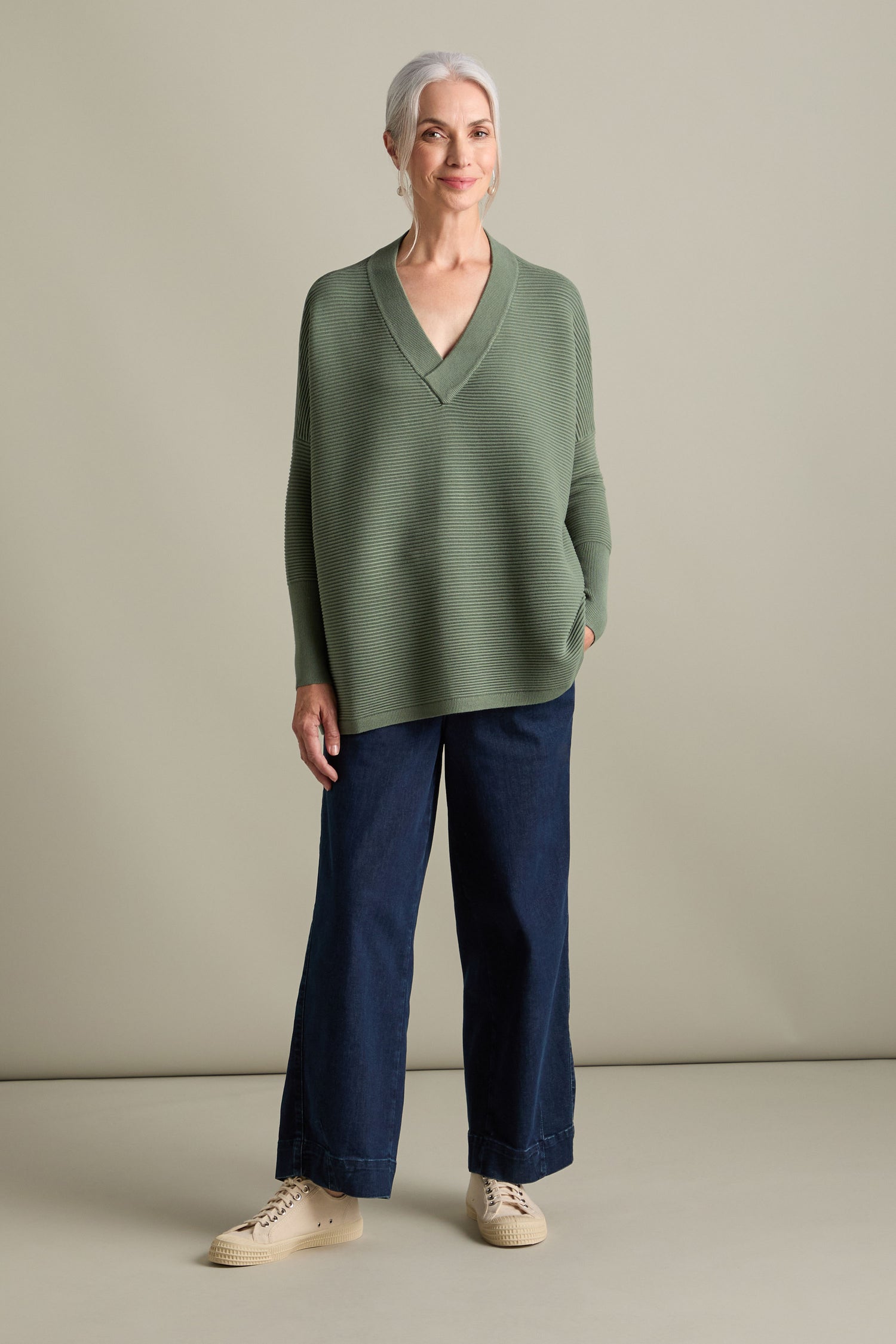 A woman with gray hair wears a V Neck Ribbed Jumper in olive green, styled in an oversized fit. She pairs it with dark blue wide-leg jeans and cream-colored shoes, standing against a plain light-colored background.