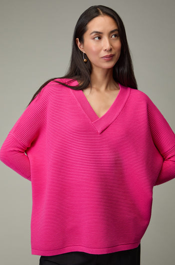 A woman with long dark hair wears the Paisie V Neck Ribbed Jumper in bright pink and black pants, standing against a plain gray background.