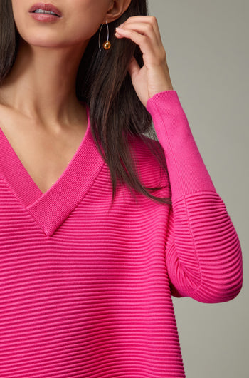 A woman poses with one hand near her face, wearing a bright pink Paisie V Neck Ribbed Jumper and gold drop earrings against a plain background.