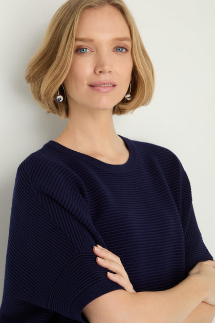 A woman with short blonde hair and dangling earrings stands with arms crossed against a plain background, wearing a Short Sleeve Navy Ribbed Top.
