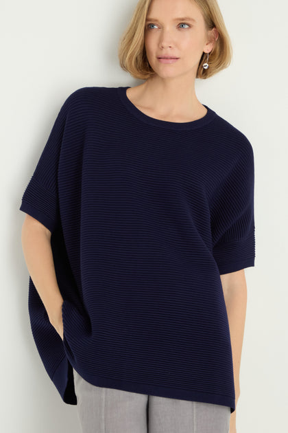 A woman wears a Short Sleeve Navy Ribbed Top paired with light gray pants, standing against a plain light-colored background.