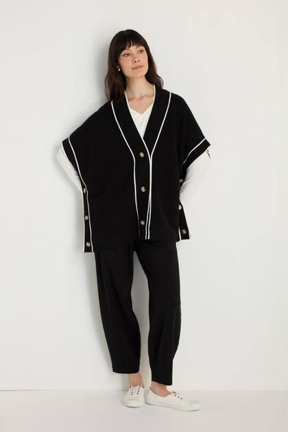 A woman stands against a white wall wearing a Piped Detail Cardigan paired with black pants, a white top, and white sneakers.