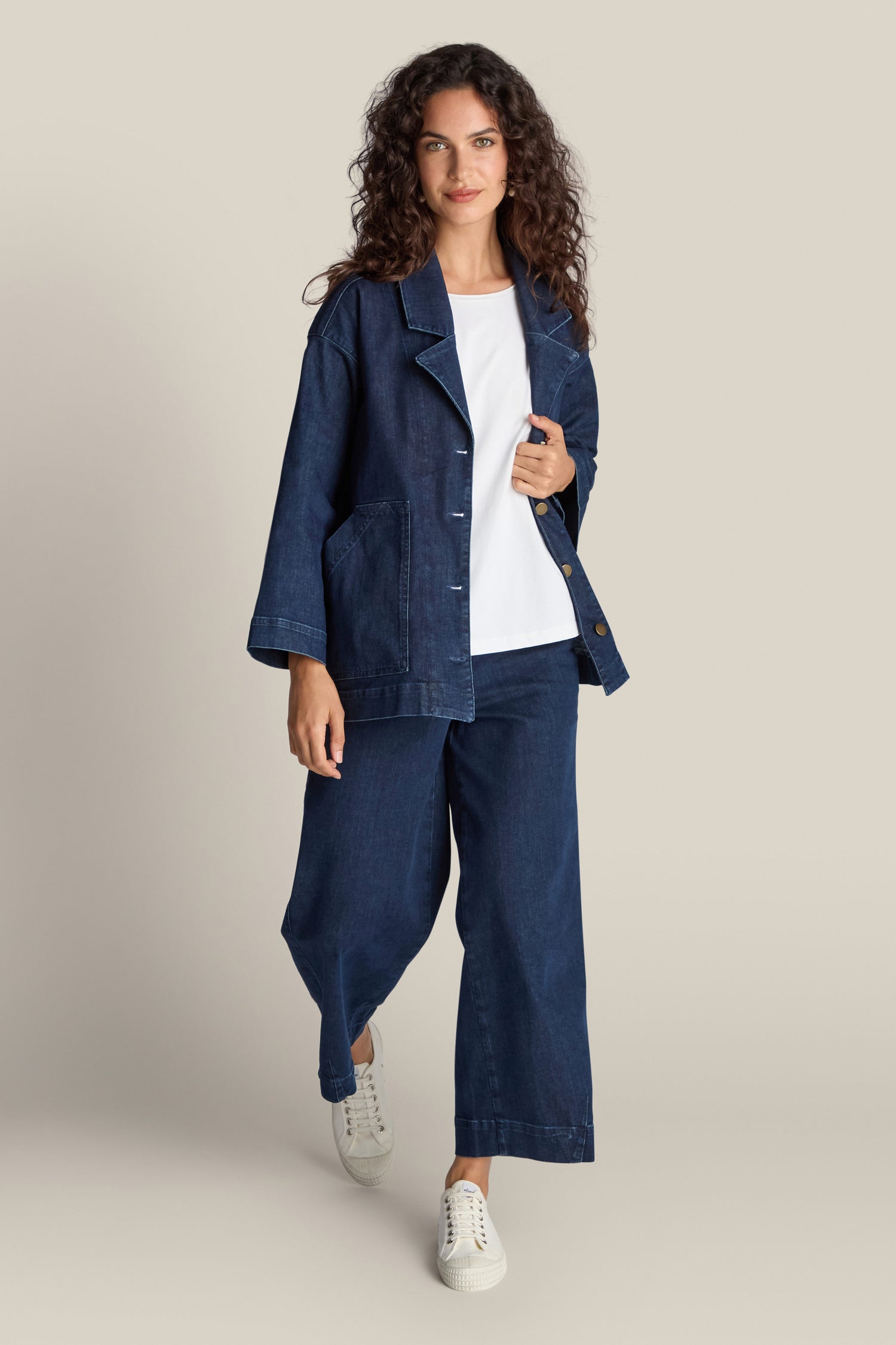 A person with curly hair is wearing the Stretch Denim Jacket, a dark blue denim piece paired with matching wide-leg pants in a relaxed silhouette, complemented by a white shirt and white sneakers, standing against a plain beige background.