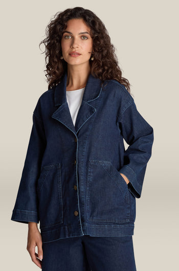 A person with long, curly hair is wearing the Stretch Denim Jacket in dark blue over a white shirt. The jacket showcases a relaxed silhouette and contemporary fit, with hands casually tucked into the pockets. The background is plain beige.