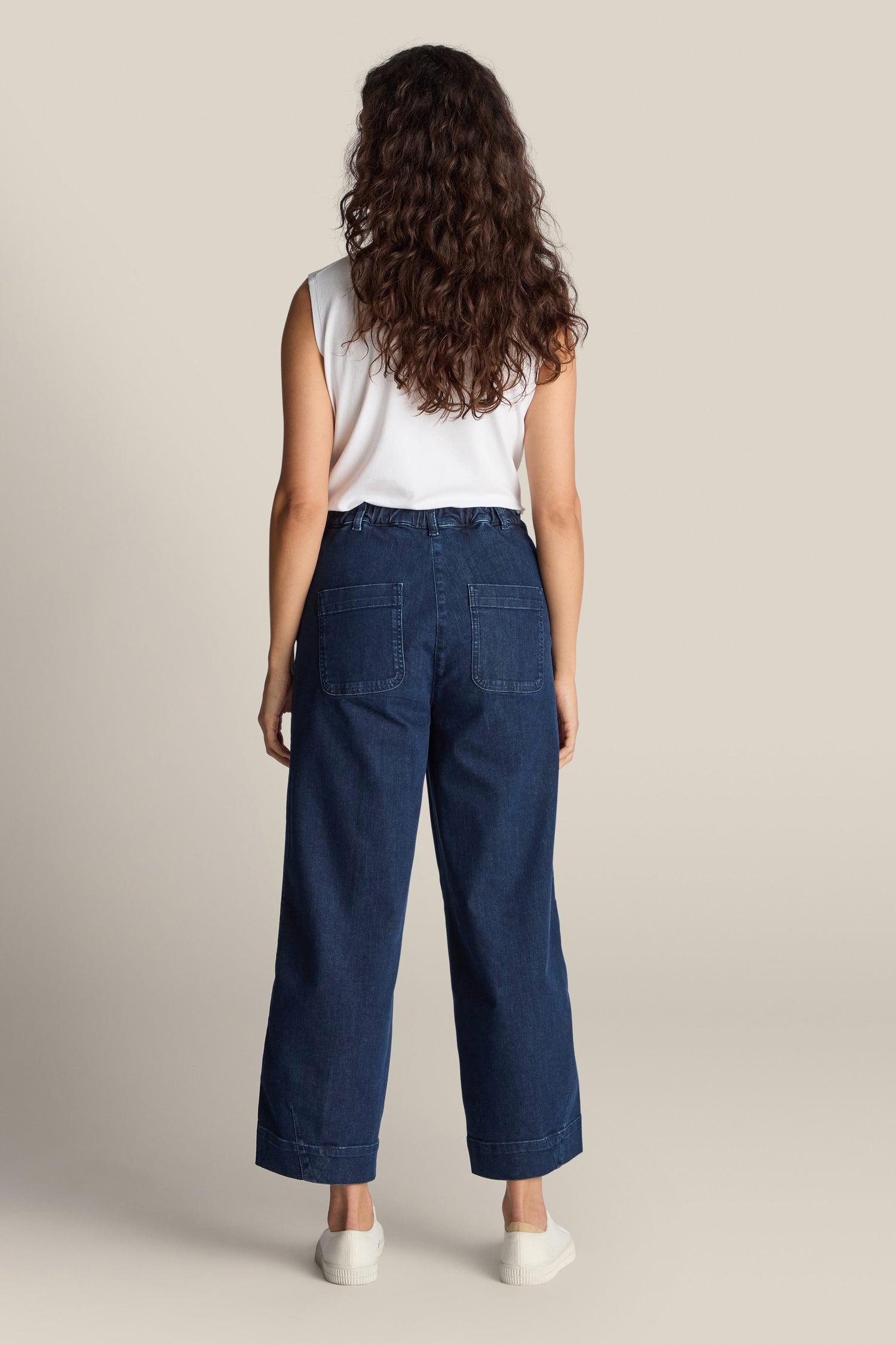 Rear view of a person with curly hair wearing a white sleeveless top, dark blue Stretch Denim Barrel Leg Trousers with an elasticated waistband, and white shoes, standing against a plain light background.