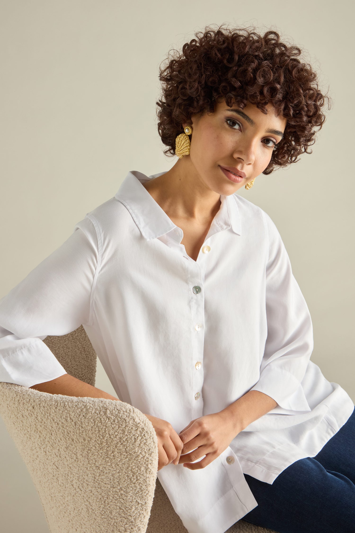 A person with curly hair, wearing a white button-up shirt and Hand Embroidered Golden Shell Glass Pearl Earrings, sits on a textured chair, slightly leaning forward.