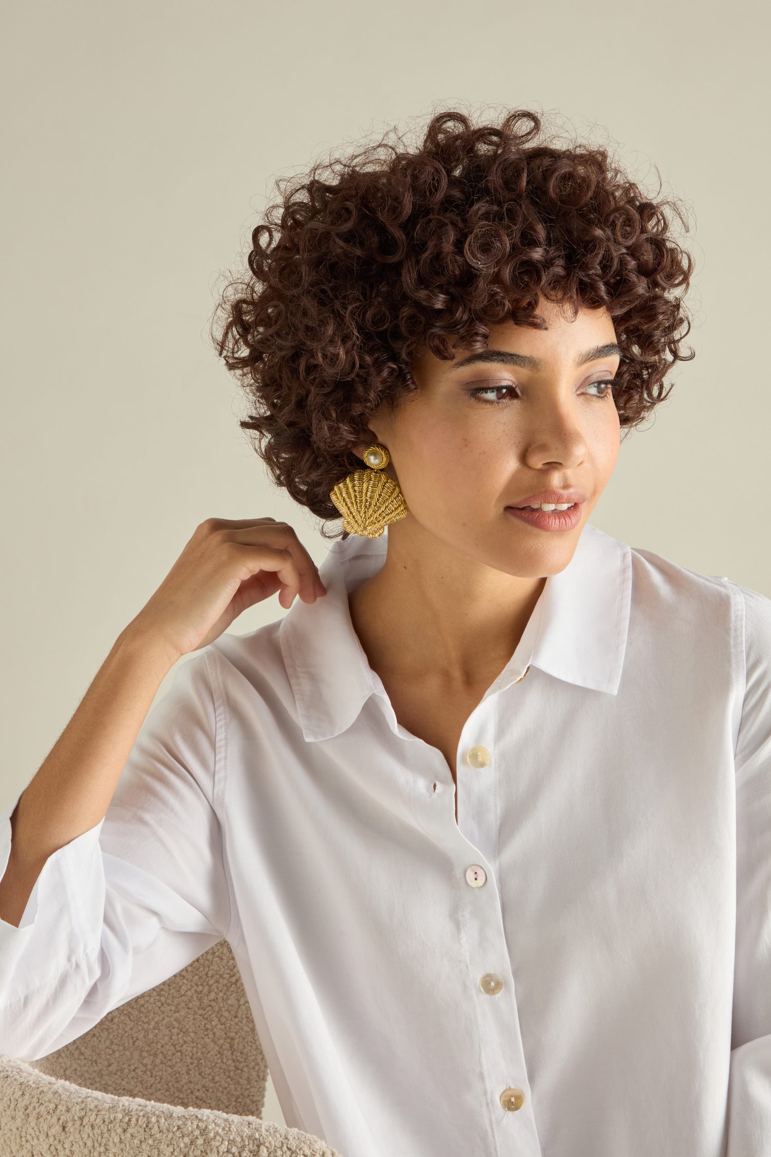 A person with curly hair in a white button-up shirt wears Hand Embroidered Golden Shell Glass Pearl Earrings, seated and looking to the side.