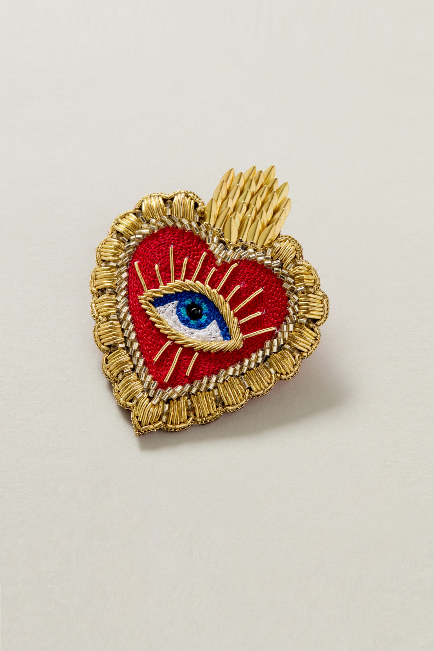 Hand Beaded Dali Heart Brooch featuring a heart shape with golden trim, an embroidered blue eye in the center, and golden rays emanating from the eye. This brooch is truly a piece of wearable art.