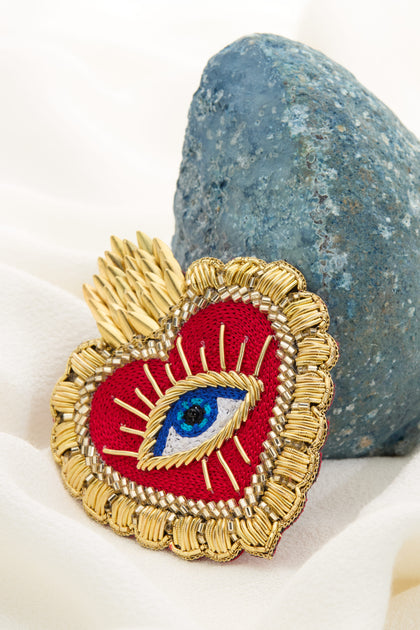 The Hand Beaded Dali Heart Brooch, adorned with a blue eye at its center, golden detailing, and wheat-like elements, rests elegantly against a textured stone on a white fabric background.