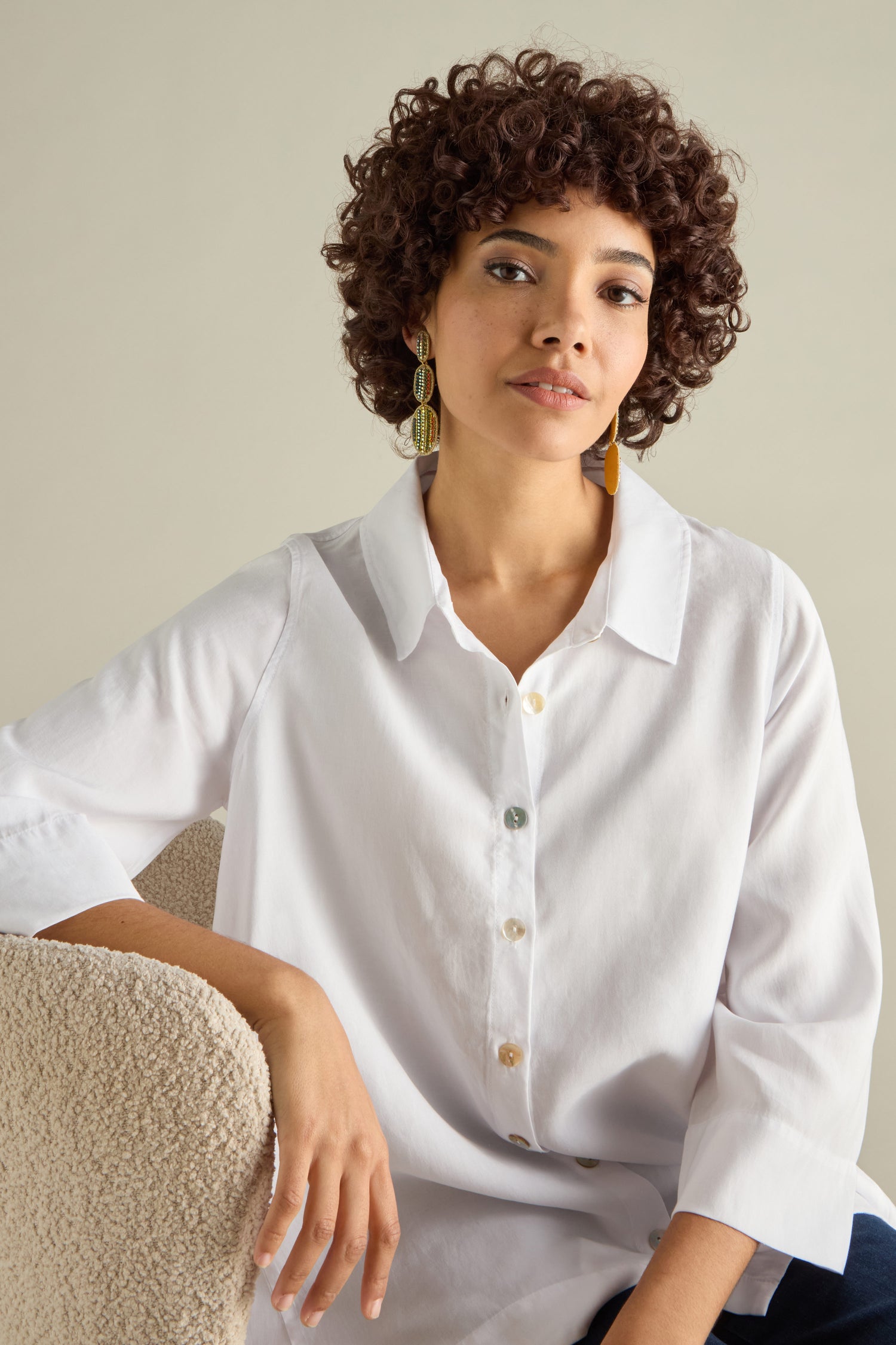 A person with curly hair sits on a chair, wearing a white button-up shirt and vibrant Hand Embroidered Crystal Bead Earrings.